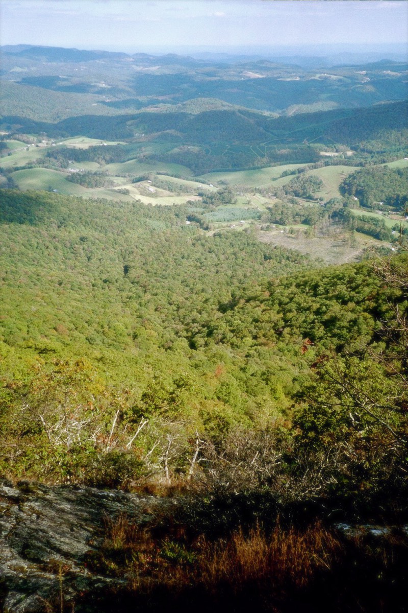 Long-range view from a High Country ridgeline overlooking forested slopes down to a distant valley of farmland